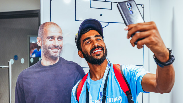 A person dressed in a Manchester City t-shirt taking a selfie with a cardboard cutout, possibly during a stadium tour.