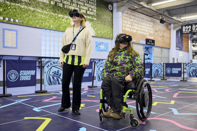 Two people experiencing a virtual reality stadium tour at Manchester City. One is standing and the other is seated in a wheelchair. Manchester City logos and stadium tour banners are visible.