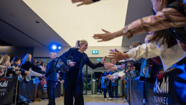 A celebrity with a blurred face interacts with fans in a tunnel-like entrance, greeting and shaking hands while holding a bag. Fans on both sides reach out, and the setting has blue lighting and branding for Asahi Tunnel Club.