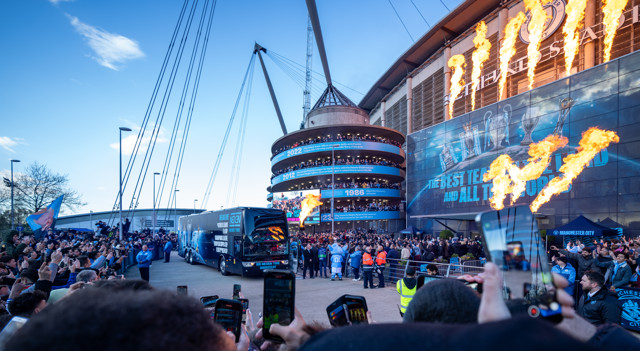 Manchester City fans outside the Etihad Stadium, watching the team bus arrive amidst a backdrop of flames and large displays, with a celebratory atmosphere in the air.
