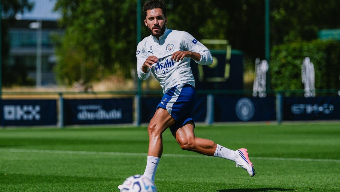 A soccer player in Manchester City training attire running on a field with a ball in front of him.