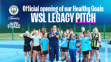 Group of young footballers and a coach standing on a blue sports pitch with Manchester City community logos. Text reads 'Official opening of our Healthy Goals WSL Legacy Pitch.'