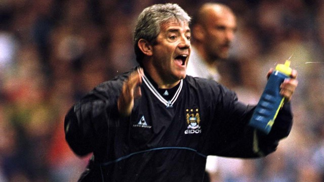 A man in a Manchester City tracksuit is animatedly gesturing on the sidelines during a football match, holding a water bottle.