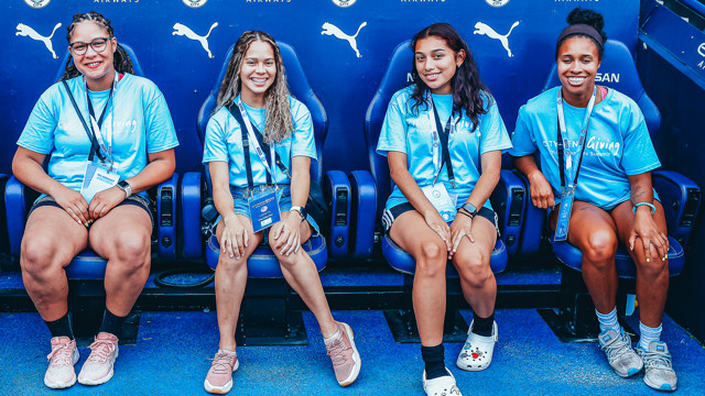 Four women, wearing blue shirts and lanyards, sit on a blue bench with the Manchester City logo and other sponsors in the background.