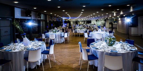 An elegant banquet hall with round tables set for a formal event. Each table features white tablecloths, blue napkins, glassware, and centerpieces with flowers. The room is well-lit with a ceiling full of small lights and wooden flooring.