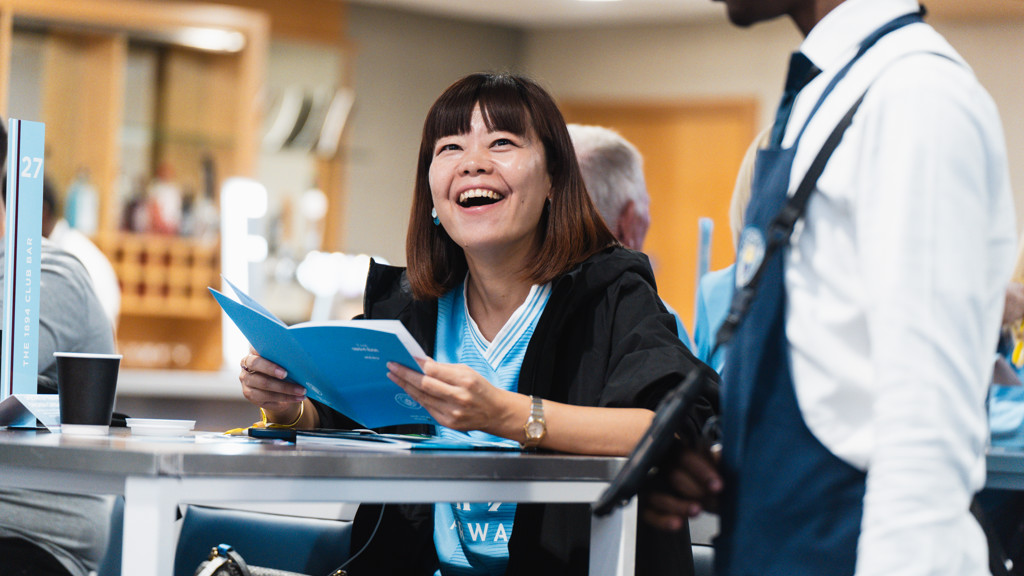 A woman in a light blue shirt is sitting and smiling, holding a brochure. She is at a table in a social setting with people around. A person in an apron stands nearby.