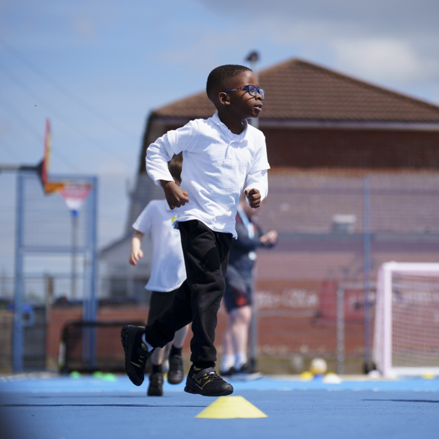 A child wearing a white shirt and black pants runs on a blue surface outdoors, with other children in the background.