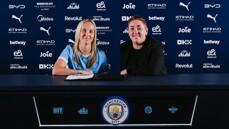 Two people sitting at a table with Manchester City branding behind them, possibly during a contract signing event.