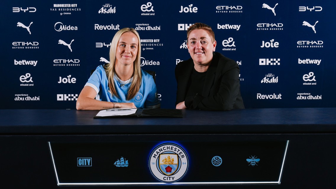 Two people sitting at a table with Manchester City branding behind them, possibly during a contract signing event.