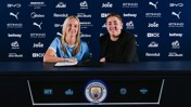 Two people sitting at a table with Manchester City branding behind them, possibly during a contract signing event.