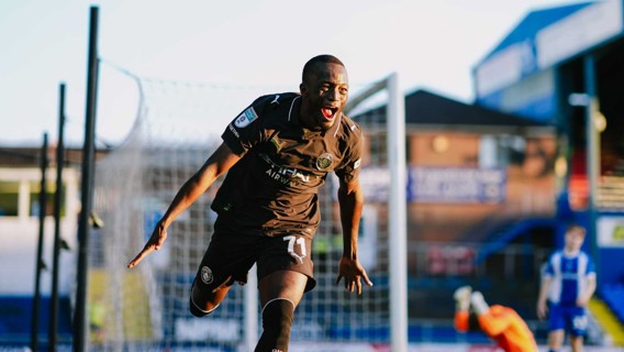 Soccer player in black jersey celebrating after scoring a goal. Stadium setting with a blurred goalkeeper in the background.