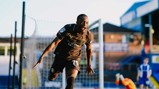 Soccer player in black jersey celebrating after scoring a goal. Stadium setting with a blurred goalkeeper in the background.