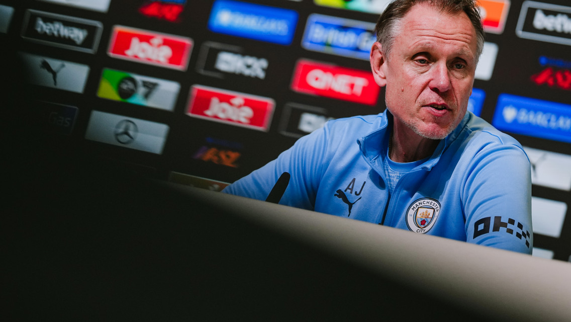 A press conference featuring a person in a Manchester City jacket in front of sponsor logos.
