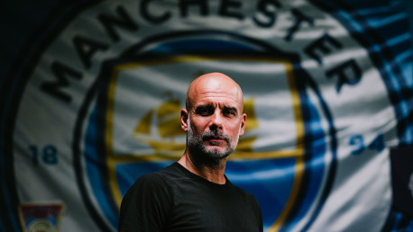 A man in a black shirt stands in front of a blurred Manchester City logo.