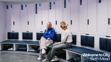 Two people sit in a Manchester City women's locker room. The lockers are labeled with names and numbers like Rose, Bunny, Greenwood, Clinton. The text 'Welcome to City Beth Mead' is visible.