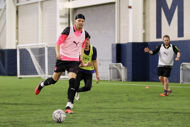 Players are training and practicing football on an indoor field wearing Manchester City training kits and equipment. There is a goal post and training equipment visible in the background.