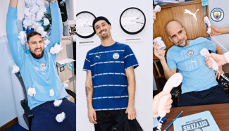 Three individuals in an office setting wearing Manchester City apparel surrounded by props like paper balls, clocks, and telephones.