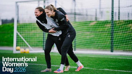 Two people in training attire engaged in a playful wrestle on a football field with 'International Women's Day 2026' text overlay.