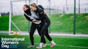 Two people in training attire engaged in a playful wrestle on a football field with 'International Women's Day 2026' text overlay.