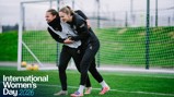 Two people in training attire engaged in a playful wrestle on a football field with 'International Women's Day 2026' text overlay.