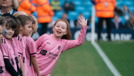 A young girl in a pink soccer uniform waves enthusiastically while standing in a line with other children before a match.