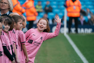 A young girl in a pink soccer uniform waves enthusiastically while standing in a line with other children before a match.