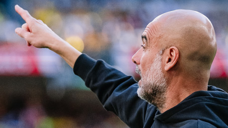 Bald man in a black jacket pointing upward in a sports venue.