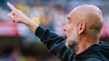 Bald man in a black jacket pointing upward in a sports venue.