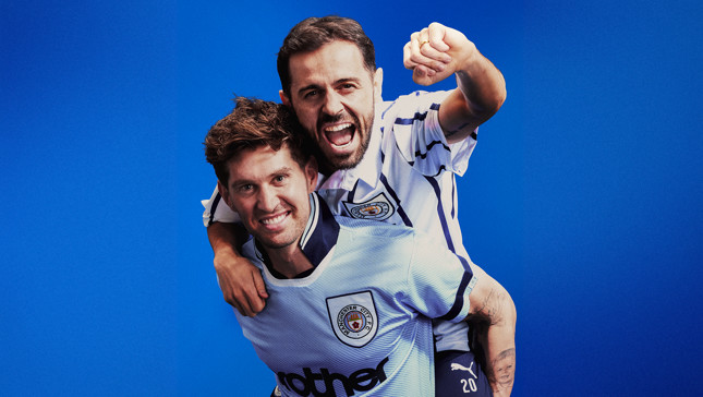 Two people in Manchester City jerseys celebrate with one carrying the other on shoulders against a blue background.