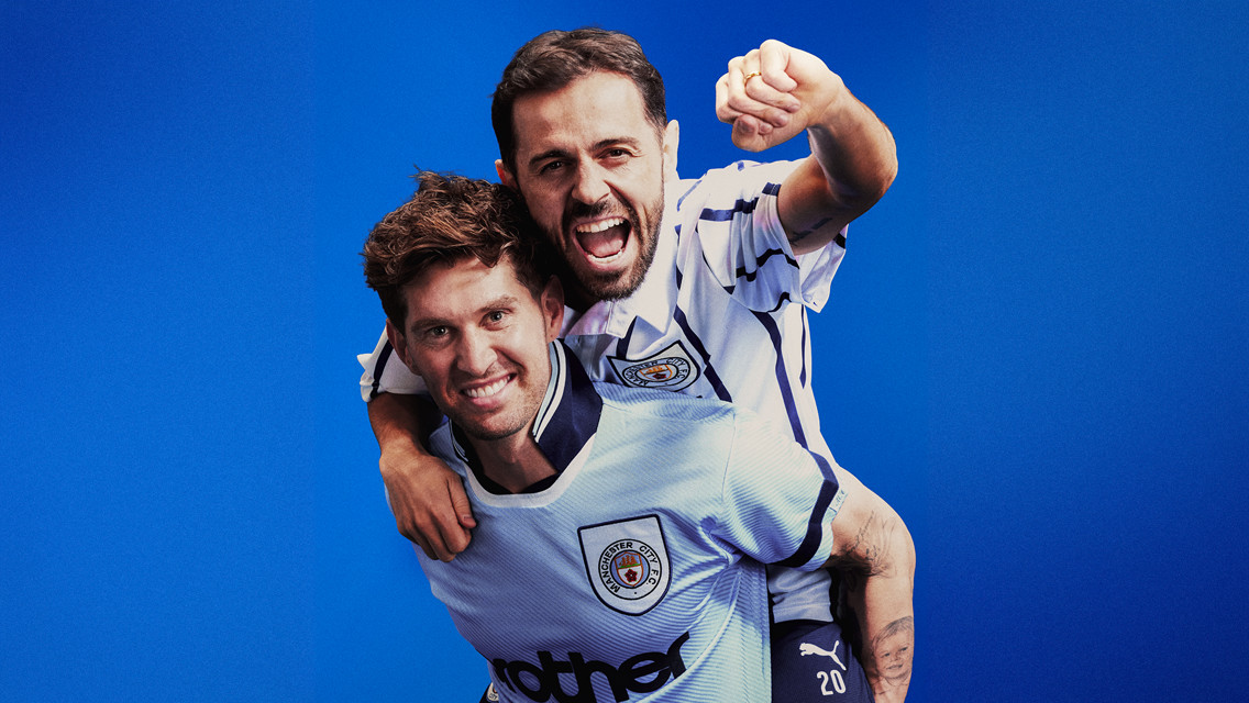 Two people in Manchester City jerseys celebrate with one carrying the other on shoulders against a blue background.