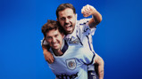 Two people in Manchester City jerseys celebrate with one carrying the other on shoulders against a blue background.