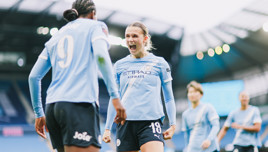 Two female soccer players in Manchester City Women's team kit celebrating a goal on the field, with a background of the stadium and other players visible.