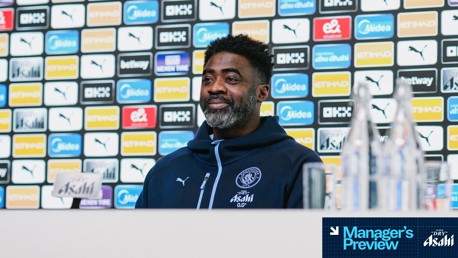 A person in Manchester City gear is at a press conference with sponsor logos in the background. The person's face is blurred.