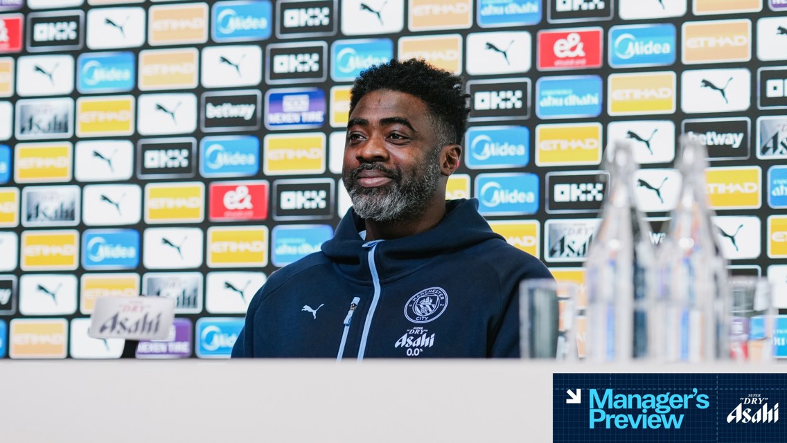 A person in Manchester City gear is at a press conference with sponsor logos in the background. The person's face is blurred.