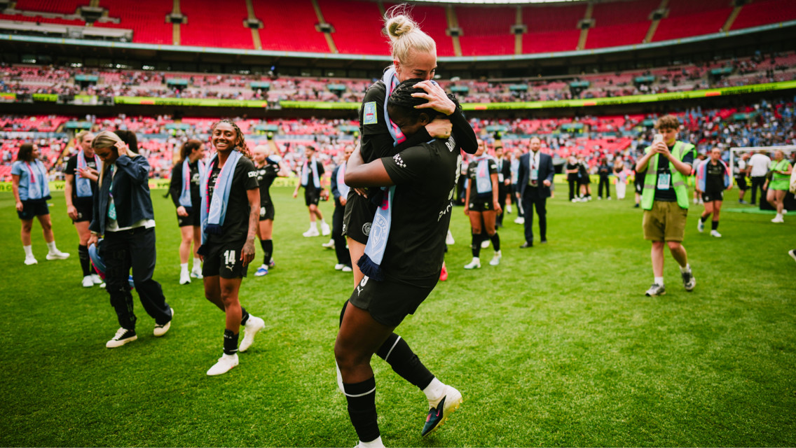 A soccer player lifts another player in celebration on a field, surrounded by other team members and staff with scarves. The stadium seats are red and there's a crowd in the background.