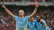 Football players in Manchester City jerseys celebrating a goal. The central player has arms raised, smiling, with teammates behind him in jubilant expressions.
