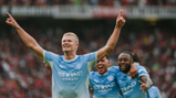 Football players in Manchester City jerseys celebrating a goal. The central player has arms raised, smiling, with teammates behind him in jubilant expressions.