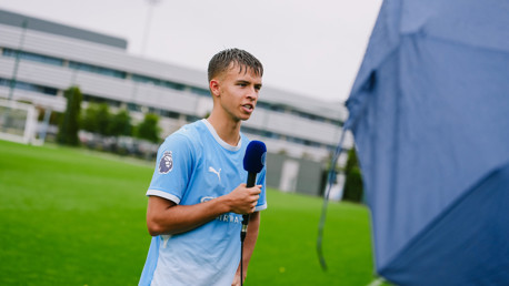 A young football player wearing a Manchester City jersey being interviewed on a soccer field. He is holding a microphone with a blue cover.