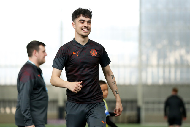 An image of a Manchester City football player during a training session. The player is wearing a Puma branded training top. Other individuals are visible in the background.