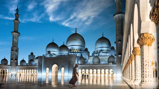 The image features the Grand Mosque beautifully illuminated at night. The blue sky and scattered stars create a serene backdrop, highlighting the mosque's domes and minarets. A person is seen walking in the mosque's courtyard.