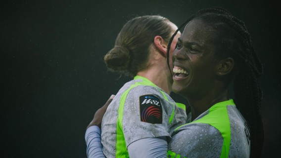 Two women soccer players in Barclays WSL jerseys embrace, with their faces blurred, showing a celebration moment on the pitch.