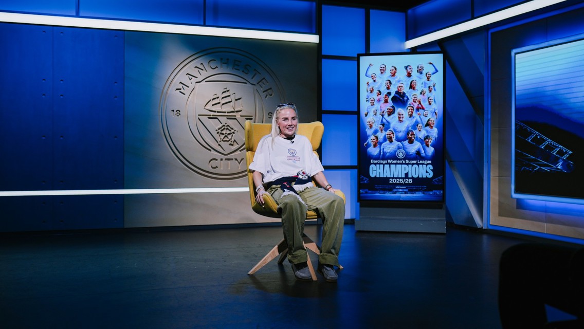A person seated in a Manchester City studio with a backdrop featuring the club logo and a poster celebrating the Women's Super League champions for the 2025/26 season.
