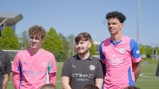 Three individuals wearing Manchester City football kits stand together on a field. They appear to be participating in a football event or gathering.