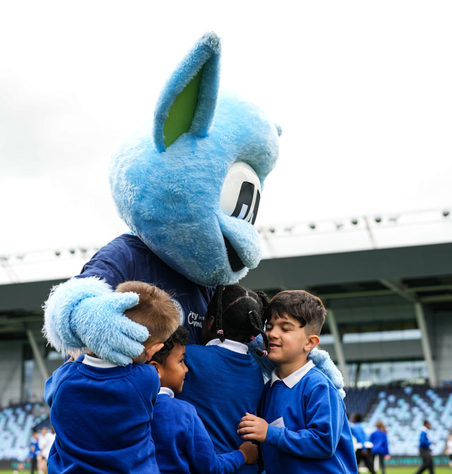 The image depicts the Manchester City mascot, Moonchester, hugging a group of school children dressed in blue uniforms at a football stadium.