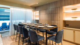 An elegant dining room with a set table for eight guests, featuring blue chairs, a wooden wall with shelves, and curtain dividing the room from blue stadium seats, suggesting a hospitality suite.