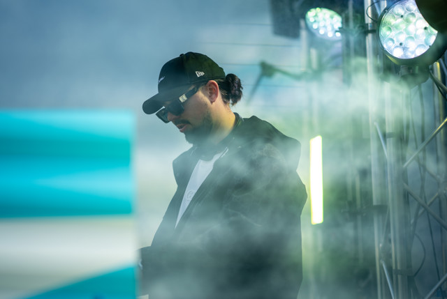 A man wearing a dark baseball cap and hooded jacket, with his face blurred, standing among stage lighting and smoke effects.