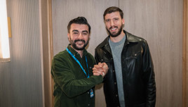 Two people shaking hands indoors, one wearing a green shirt with a lanyard and the other wearing a black leather jacket.