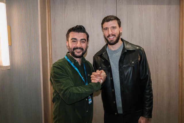 Two people shaking hands indoors, one wearing a green shirt with a lanyard and the other wearing a black leather jacket.