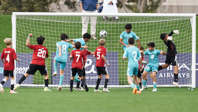A group of young soccer players in red and blue uniforms are attempting to score a goal during a match. The goalkeeper is making a dive to stop the ball, with spectators watching in the background.