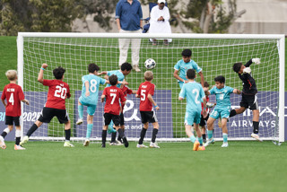 A group of young soccer players in red and blue uniforms are attempting to score a goal during a match. The goalkeeper is making a dive to stop the ball, with spectators watching in the background.